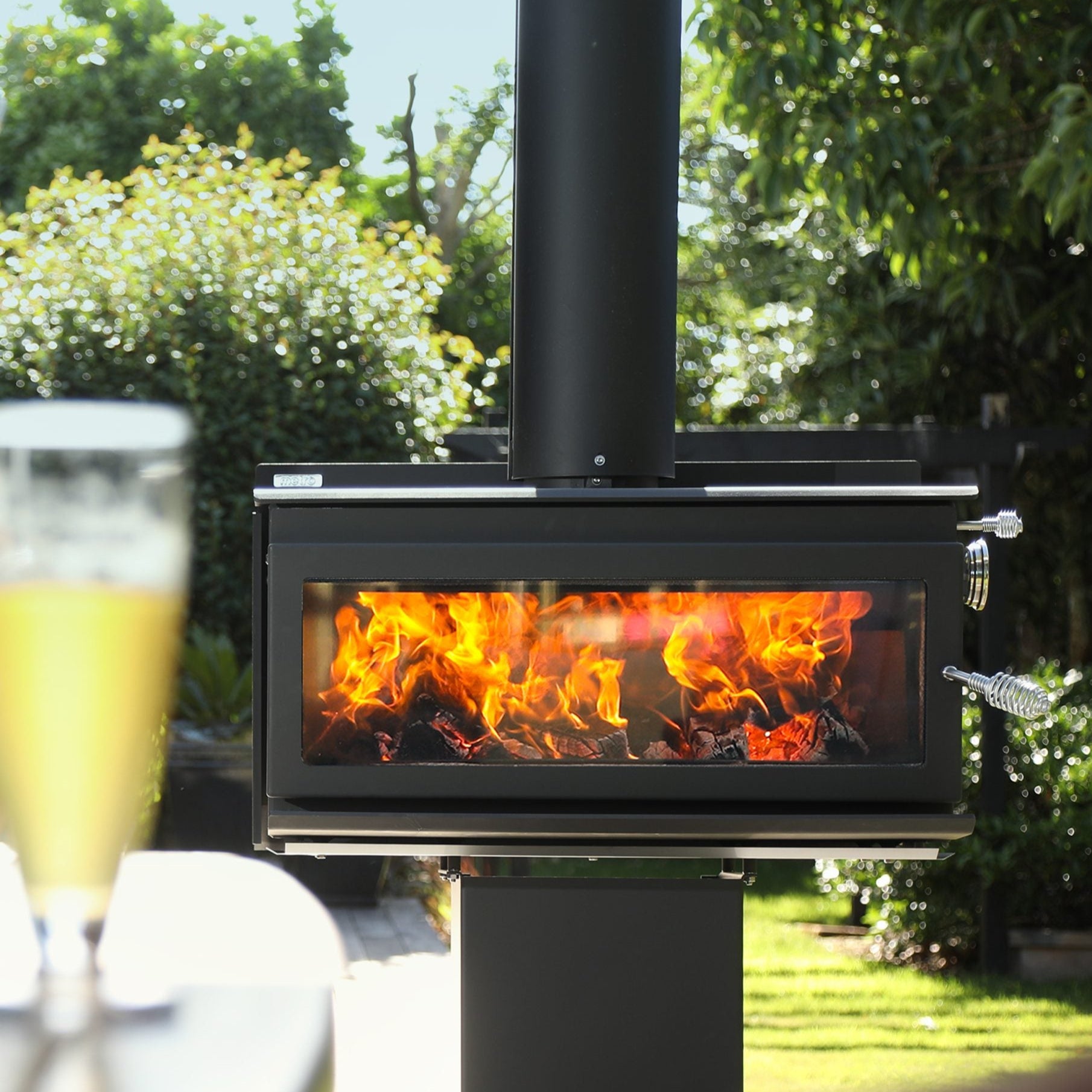 An outdoor wood-burning fireplace with a black Corten steel pedestal base, featuring a visible fire through the glass door, set in an outdoor setting.
