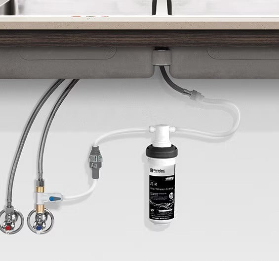 An undersink water filtration system installed under a kitchen bench, consisting of a cylindrical cartridge filter and connected tubing.